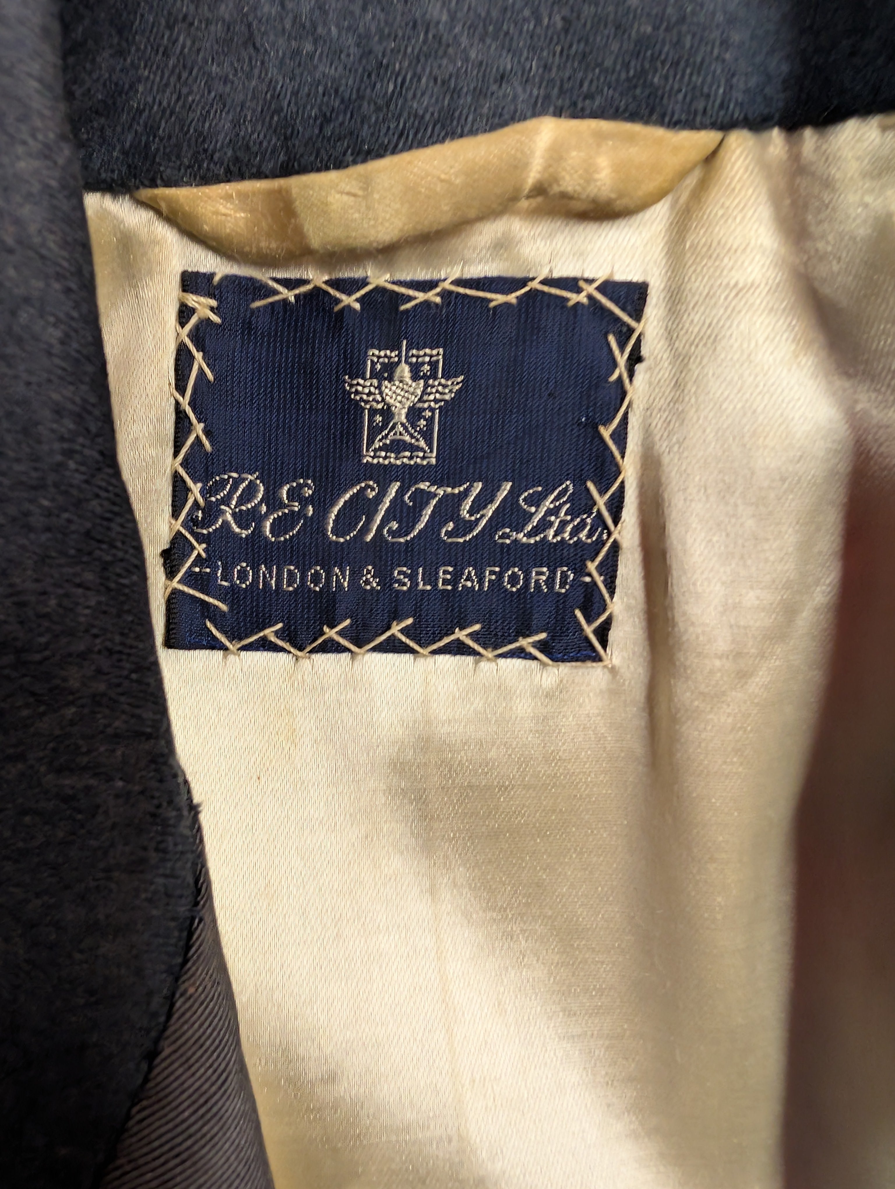 Close-up of the interior neck label of a dark gray or charcoal-colored jacket. The label is navy blue with gold embroidered text reading 'RE CITY Ltd LONDON & SLEAFORD' and a gold crest, set against a cream-colored lining.