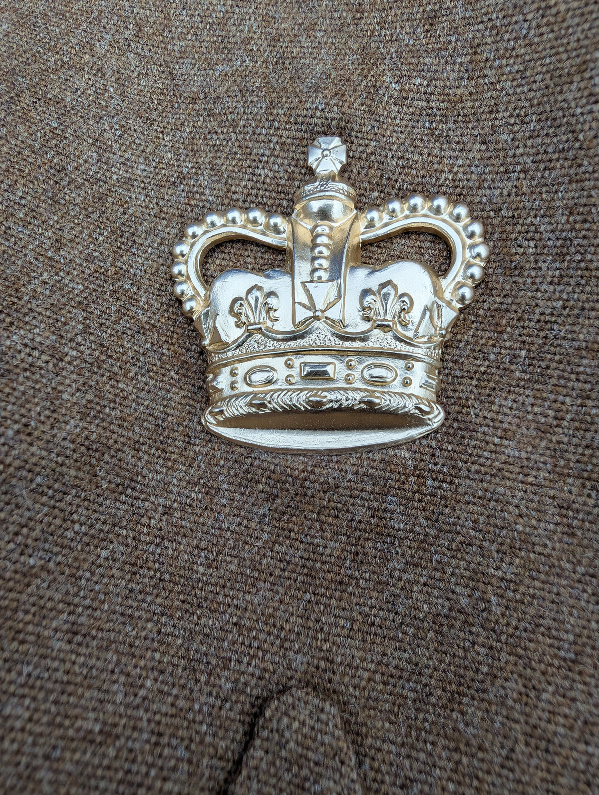 A silver-toned metal crown-shaped brooch or pin with ornate detailing and a small clear stone, photographed on a brown textured fabric background.