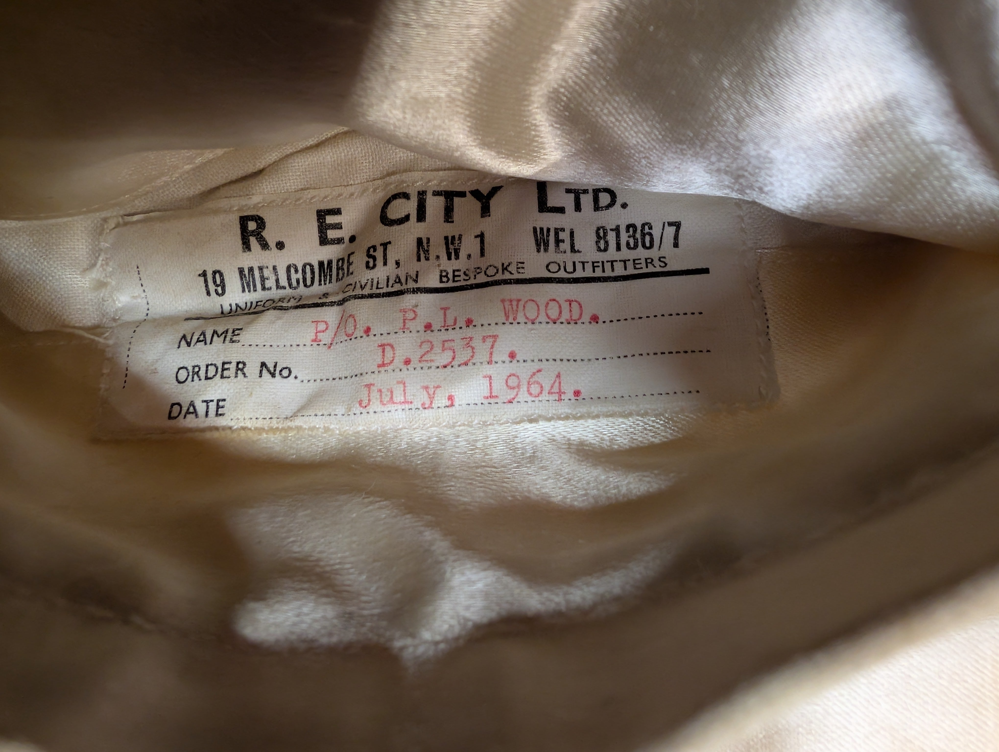 Close-up of an interior fabric label sewn onto a light beige or cream garment. The label reads R.E.City Ltd Tailors London Seaford with P/O RAF Officer P.L.Wood Dated July 1964, RAF Service Number D.2537