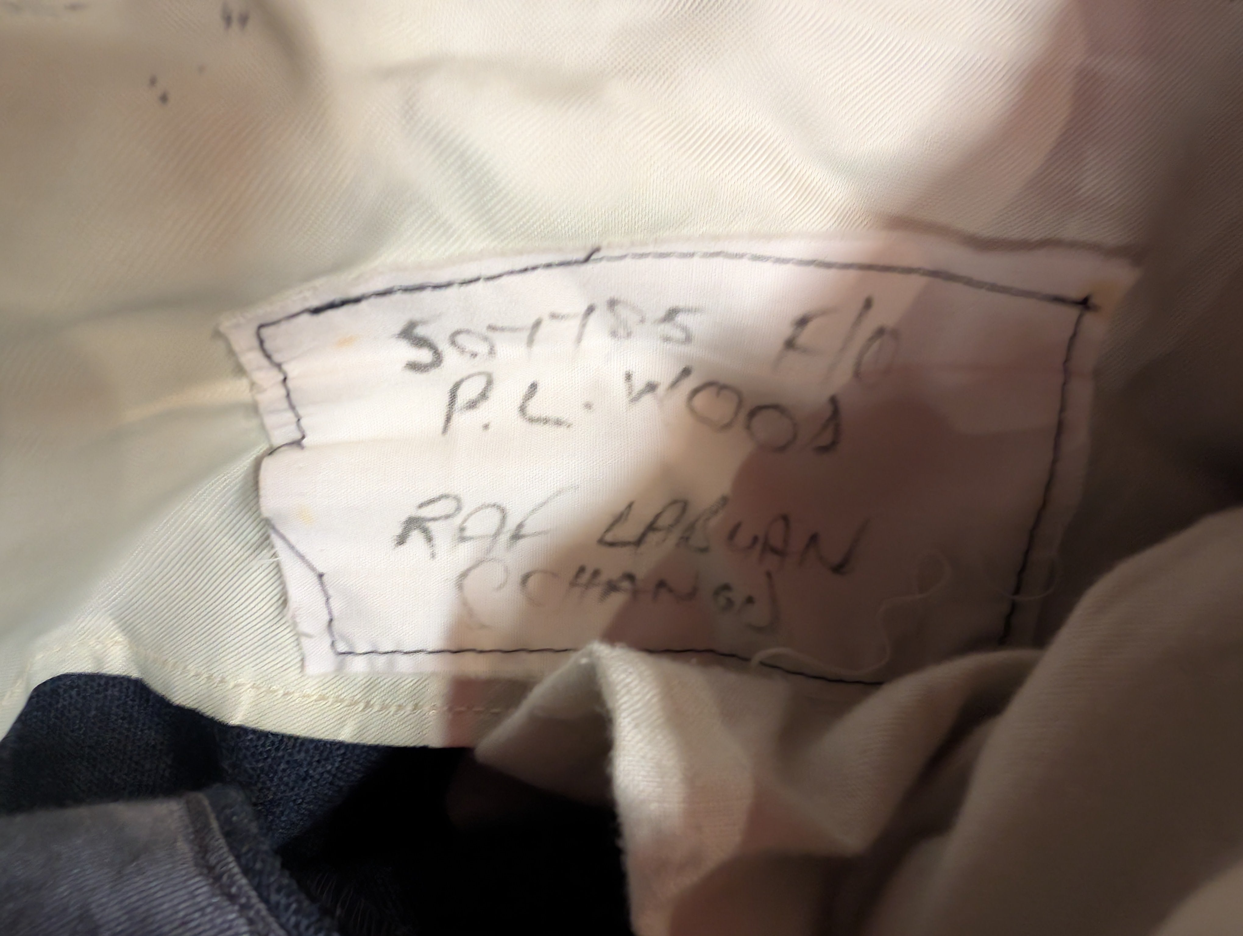 A close-up of a sewn-in fabric label inside a garment, with handwritten text reading '507785  P.L.WOOD, RAF Labuan